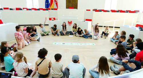 Kinder sitzen in einem großen Kreis auf dem Fußboden des Plenarsaal. In der Mitte liegen Bilder.