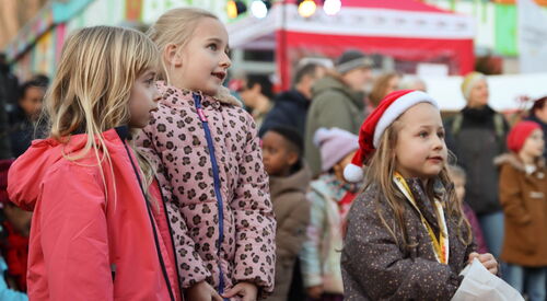 3 staunende Mädchen im Vordergrund. Das Mädchen rechts trägt eine rote Weihnachtsmütze. Im Hintergrund weitere Gäste.