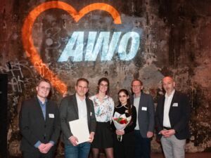 Sechs Personen. Vier Männer, zwei Frauen. Vor einer Wand mit AWO-Logo. Festlich gekleidet. Eine Frau hält einen Blumenstrauß in den Händen.