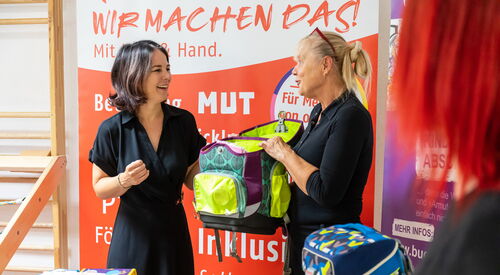 Zwei Frauen sehr fröhlich halten einen Schulranzen zwischen sich. Auf einem Tisch vor ihnen liegen mehrere bunte Federmappen für Kinder. Ein blauer Kanzen steh auf auf dem Tisch.