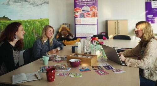 Beratungsraum des Büro Kindermut mit lila Roll-Up in der linken hinteren Ecke. Drei Frauen sitzen am Tisch. Zwei auf der linken Seite nebeneinander, sie lächeln. Die andre gegenüber mit Laptop vor sich.