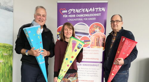 Zwei Männer und eine Frau halten große Zuckertüten in den Händen und lächeln. Sie stehen vor einen lila Aufstteller vom Büro Kindermut.