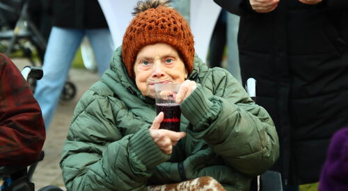 Alte Frau im Rollstuhl. Sie ist dick und warm angezogen mit Decke über den Beinen. Sie hat ein Getränk in den Händen.