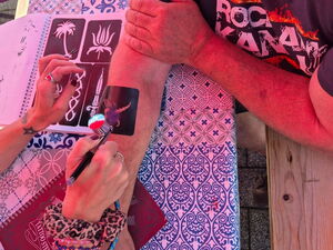 Auf einem Unterarm wird mit einer Schablone und Farbe ein Tattoo-Motiv aufgesprüht. Daneben liegt eine Vorlage mit weiteren Motiven.