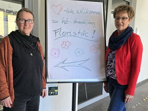Zwei Frauen stehen im Flur, zwischen ihnen ein Flipchart mit Wegweiser-Pfeil zur Info-Veranstaltung Floristik.