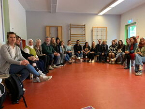 Gruppenfoto der Teilnehmenden. Sitzen in großem Halbkreis in einer Turnhalle/Sportraum.