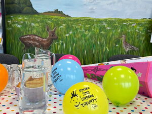 Auf einem Tisch: bunte Luftballons und Zuckertüten. Dahinter das Bild einer Wiese mit Rehkitz und Vögel.
