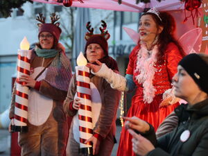2 Frauen in Rentier-Kostümen halten große Kerzen-Atrappen. Daneben steht Fee Lilli im roten Kleid, mit zarten Flügeln und Krone. Vorne rechts Gebärdendolmetscherin.