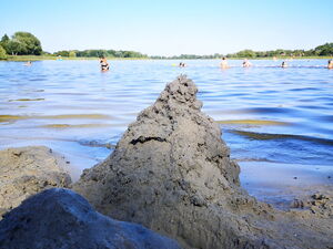 Eine Sandburg am Ufer eines Sees. Dahinter Wasser, blauer Himmel und am Horizont Baumreihen.