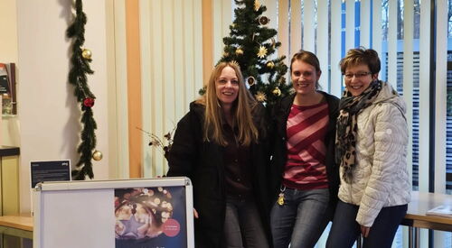 Weihnachtlich geschmückter Raum. 3 Frauen lehnen sich an einen Tisch. Sie lächeln in die Kamera.