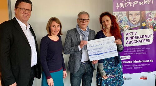 4 Personen stehen nebeneinander. 2 von ihnen halten eine Check hoch, rechts daneben das Plakat des Büro Kindermut.