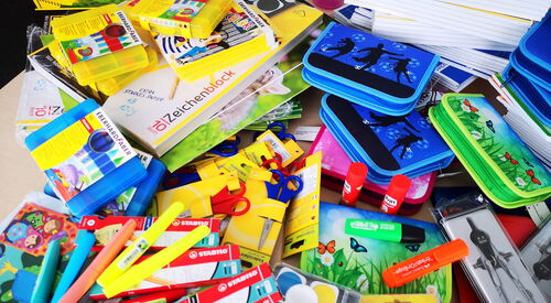 Verschiedene Schulmaterialien wie Stifte, Wasserfarbenkästen, Lineale, Dreieck, Zirkel, Spitzer und bunte Federmappen, durcheinander auf einem Tisch verteilt.