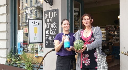 2 Frauen auf den Stufen zu einem Geschäft. Frau rechts hält Spendendose, Frau links hat eine Melone in beiden Händen. Beide lächeln.