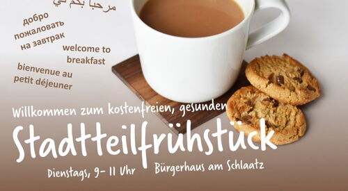 Eine weiße Tasse mit Kaffee steht auf einem Holztisch neben zwei Schokokeksen. Im Hintergrund ist Text mit der Einladung zum „Stadtteilfrühstück“ im Bürgerhaus am Schlaatz.