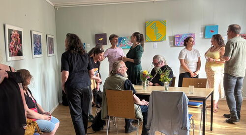 Ausstellung. Bilder hängen an weißen Wänden. Menschen sehen sich die Bilder an.