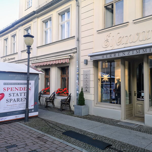 Ladengeschäft in Einkaufsstraße. Herrenzimmer. Links davon eine Laterne und eine kleine Bude mit dem Banner: Spenden statt sparen an der Außenwand.