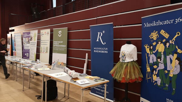 Informationsstände mit Roll-ups und Broschüren entlang einer Wand in einem Veranstaltungsraum.