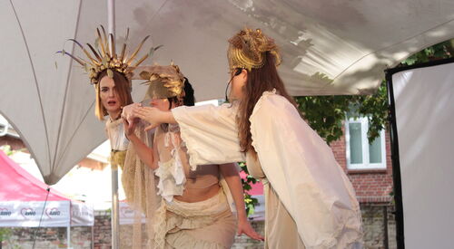 Schauspiel. 3 Frauen in weißen / beigen Kleidern / Gewändern mit Schmuckvollen Haarschmuck ähnlich Kronen. Von der Seite.