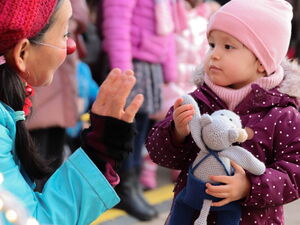 Kleines Kind steht einer Clownin gegenüber die winkt, Kind hält ein Kuscheltier in den Händen.