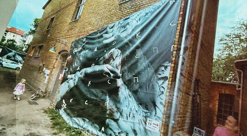 Schief-verzerrtes Foto. Eine Hauswand mit einem düsteren Bild auf großem Tuch. Ein kleines Kind läft allein und dem großwirkenden Haus vorbei.