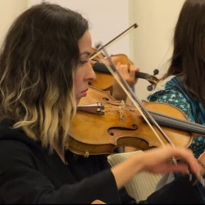 Nahaufnahme. Orchester. Mehrere Personen spielen Geige. Im Vordergrund zwei Frauen.
