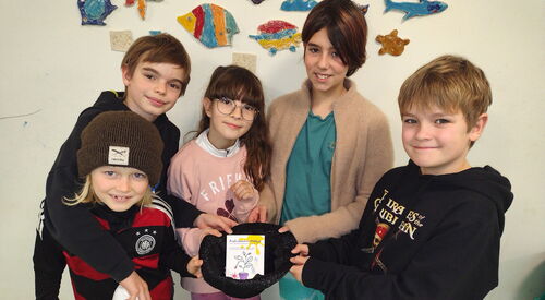 5 Kinder eng beieinander halten eine Mütze in die Mitte, darin eine Karte. Die Kinder sind fröhlich.