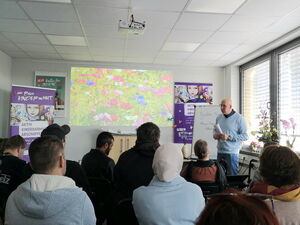 Großer Raum. Teilnehmende sitzen in Stuhlreihen, vorne steht ein Mann rechts am Fenster.