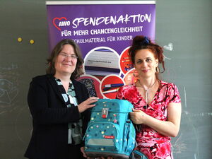 2 Frauen halten ein türkisen Schulranzen zwischen sich und lächeln. Hinter ihnen ein lila Roll-Up Spendenaktion.