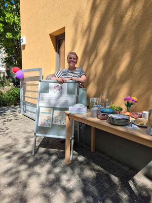 Sonniger Tag, Büfett links daneben ein Aufsteller mit mehreren Informationen, eine Frau steht dahinter und lächelt in die Kamera.