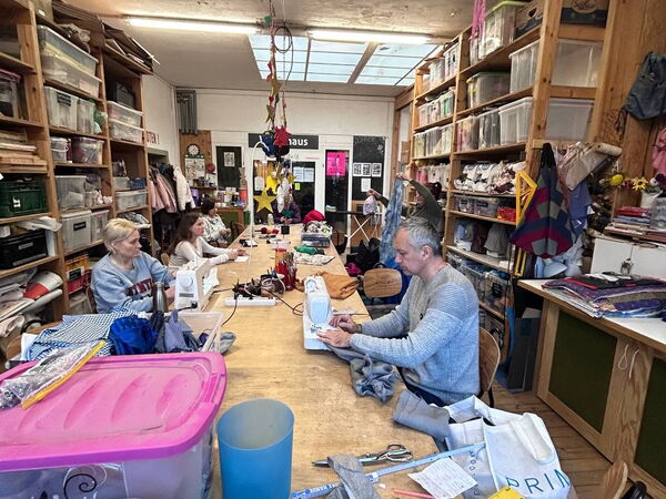 Material, Stoffe im Vordergrund. Menschen an langem Tisch vor sich Nähmaschinen.