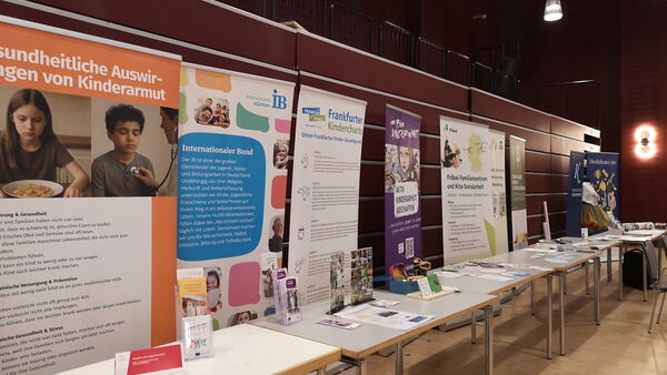 Lange Tischreihe mit aufgestellten Informationsplakaten und Broschüren in einem Veranstaltungsraum. Armut bekämpfende Einrichtungen stellen sich vor.