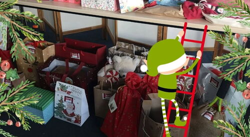 Bearbeitetes Foto. Viele Geschenktüten auf dem Fußboden und auf einem Tisch direkt darüber. Bearbeitung: Ein kleine Wichtel in grüner Kleidung mit grüner Weihnachtsmütze hat eine rote Leiter angelehnt und klettert zu den Tüten.