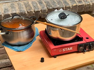 Tisch draußen. Ein Topf mit Essen auf einem Handtusch und ein Topf auf mobiler Heizplatte. Roter Campingkocher.