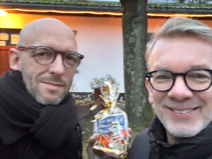 Zwei Männer stehen draußen vor einem beleuchteten Gebäude und lächeln in die Kamera, einer hält ein kleines Geschenktütchen in der Hand.