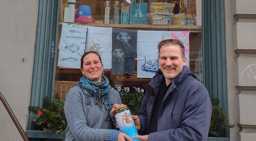 Eine Frau übergibt Spendendose an einen Mann. Beide lächeln in die Kamera. Sie stehen vor einen erleuchteten Schaufenster.