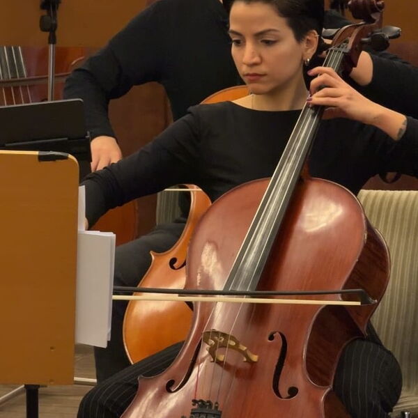 Frau in schwarzer Kleidung sitzt und spiel Cello. Sie hat kurze schwarze Haare.