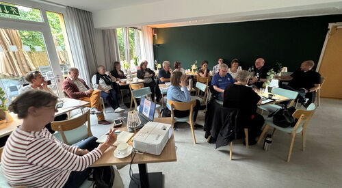 Sitzung: Größerer Raum, Menschen sitzen locker an Tischen.  Vor im Bild eine Frau an einem Tisch mit Laptop und Beamer.
