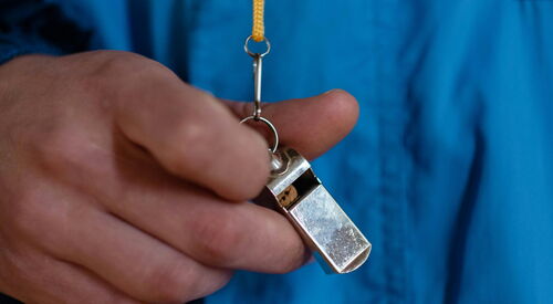 Person mit blauer Jacke und Trillerpfeife in der Hand. Nahaufnahme Trillerpfeife.