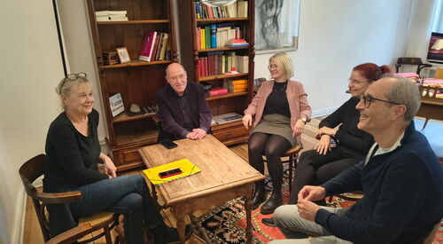 3 Frauen, 2 Männer sitzen in einem gemütlichen Raum mit Bücherregal um einen Holztisch.