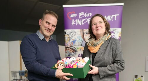 Mann und Frau stehen vor dem Rollup Banner von Büro kindermus und halten einen Karton mit Kuscheltieren.