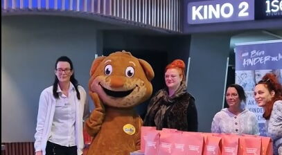Kino. Vor Kino 2 stteht ein Maskottchen und 4 Frauen. Ein DM Stand mit Geschenktüten darauf.   Text im Bild: Was ist denn hier los?