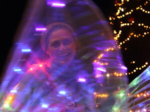 Großer beleuchteter Weihnachtsbaum. Davor Frau die lacht und ihre farbig leuchtenden Flügel bewegt.