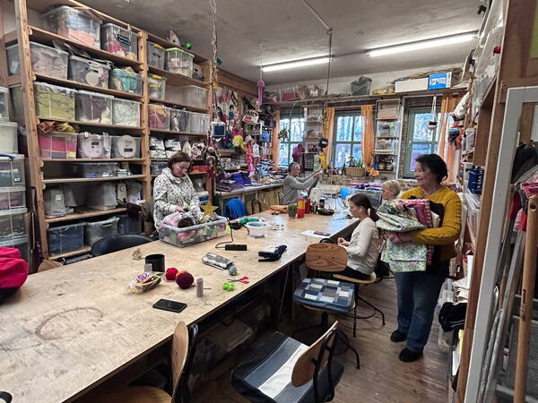 Großer Raum, Langer Tisch, viele Stühle, große Regale mit Stoff und anderen Nähutensilien. Einige Frauen am Tisch beschäftigt.