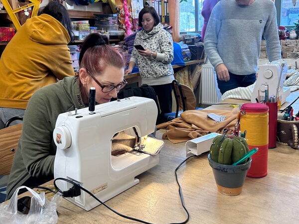 Frau sitzt an Nähmaschine. 3 weitere Personen im Raum.