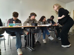 4 Jugendliche sitzen an aneinandergereihten Tischen nebeneinander. Auf den Tischen Schulmaterial. Ihnen gegenüber auf der innenseite des Tisches eine blonde Jugendliche stehend.