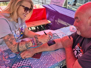 Blonde Frau mit Sonnenbrille und Tattoos auf dem Arm, sprüht/malt ein Bild mit Schablone auf den Unterarm eines älteren Mannes.