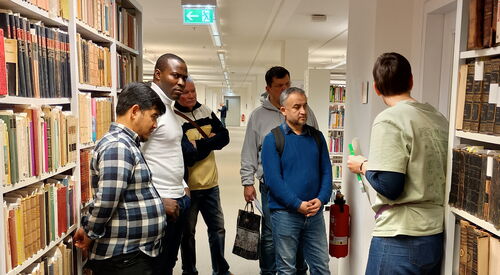 Teilnehmer werden durch die Bibliothek geführt. Sie stehen zwischen weißen Ragalen mit alten Büchern.