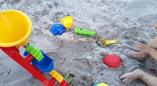Bunte Sandspielzeuge liegen im Sand, darunter ein roter Trichterturm, Förmchen und Schaufeln. Rechts sind Kinderfüße im Sand.