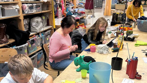 Ein Junge, eine Frau und ein Mädchen sitzen am Tisch und nähen und basteln. Hinter ihnen große gefüllte Regale.