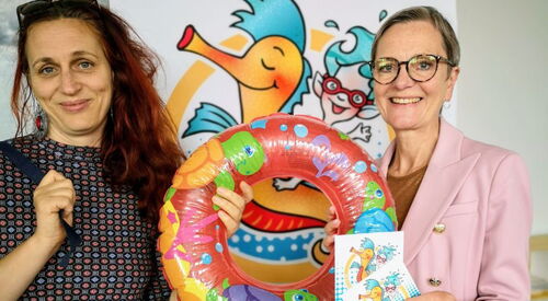Zwei Frauen mit einem bunten Kinder-Schwimmreifen zwischen sich stehen vor dem Aufsteller des Projektes Wellenreiter